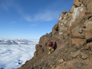 Dr Steven Andrews inspecting the boundary between successive lava flows in Wollaston Forland in northeast Greenland. The faint red band directly above the geologists head represents the eruption surface of a lava flow that was broken down into laterite by the hot wet climate.